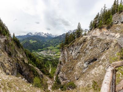 A panoramic view of a high-altitude mountain pass with wooden walkways and railings built into steep rocky cliffs, overlooking a green valley and distant snow-capped peaks.