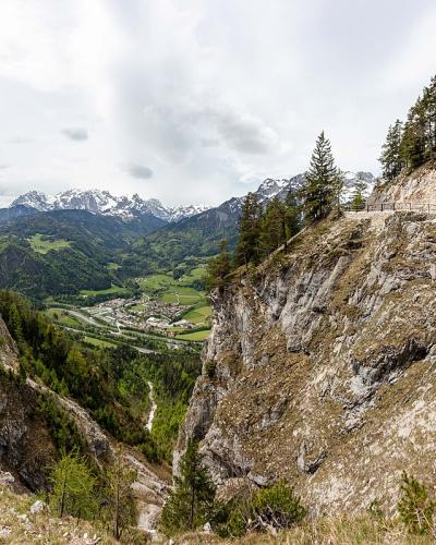 A panoramic view of a high-altitude mountain pass with wooden walkways and railings built into steep rocky cliffs, overlooking a green valley and distant snow-capped peaks.