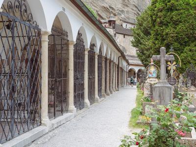A long, arched colonnade with iron-grated crypts lines a gravel path in a cemetery, with a large rock face and a small tower visible in the background.