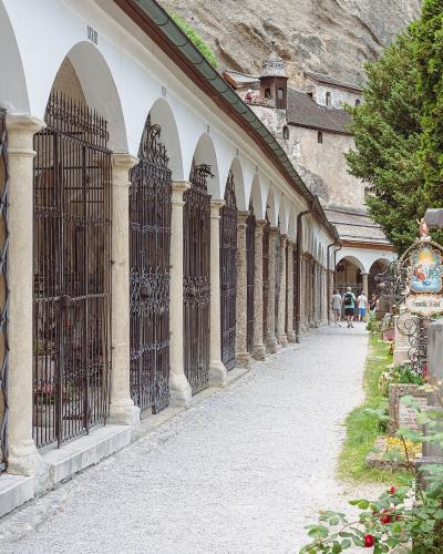 A long, arched colonnade with iron-grated crypts lines a gravel path in a cemetery, with a large rock face and a small tower visible in the background.
