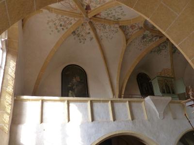The image shows the interior of a church featuring a gallery with vaulted ceilings decorated with floral motifs, religious paintings, and a small organ.