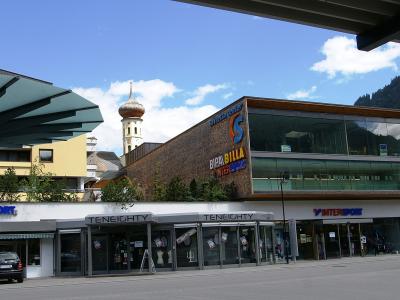 A modern commercial building complex featuring an Intersport store, a Bipa/Billa supermarket, and a Teneighty shop, with a church tower visible in the background.