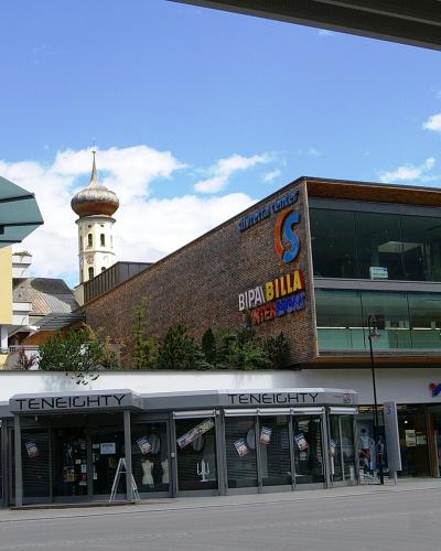A modern commercial building complex featuring an Intersport store, a Bipa/Billa supermarket, and a Teneighty shop, with a church tower visible in the background.