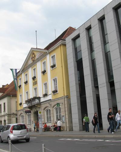 A street scene showing a yellow building with "RATHAUS" written on its facade, situated next to a modern grey building, with cars and pedestrians on the street.