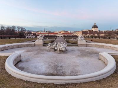 A wide-angle view of the formal Baroque gardens of the Belvedere Palace in Vienna, featuring a large, empty, kidney-shaped fountain basin in the foreground with white marble statues, leading up a central axis toward the Upper Belvedere palace and the
