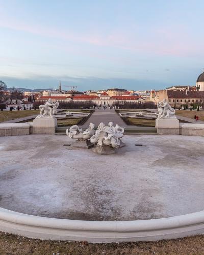 A wide-angle view of the formal Baroque gardens of the Belvedere Palace in Vienna, featuring a large, empty, kidney-shaped fountain basin in the foreground with white marble statues, leading up a central axis toward the Upper Belvedere palace and the