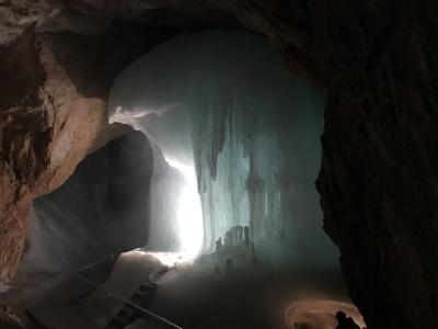 A large, illuminated ice formation inside a dark limestone cave, featuring a prominent vertical ice wall with stalactites and a metal staircase with a railing in the foreground.