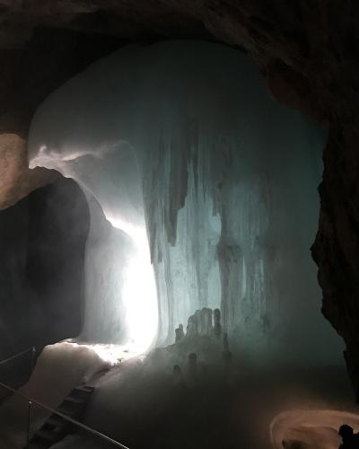 A large, illuminated ice formation inside a dark limestone cave, featuring a prominent vertical ice wall with stalactites and a metal staircase with a railing in the foreground.
