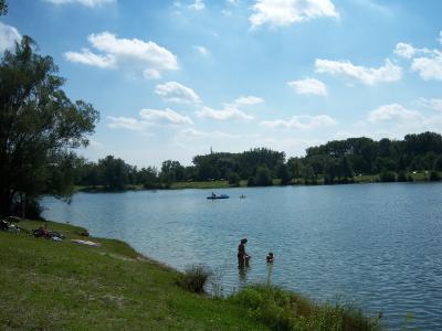 A scenic view of a large recreational lake with people swimming and boating, surrounded by green grassy banks and trees under a blue sky with scattered clouds.