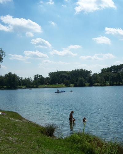 A scenic view of a large recreational lake with people swimming and boating, surrounded by green grassy banks and trees under a blue sky with scattered clouds.