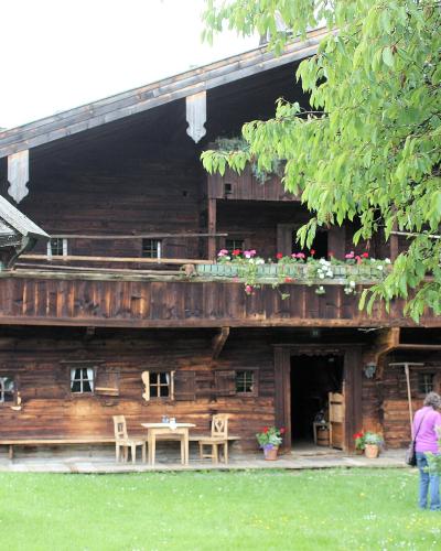 A traditional wooden Alpine farmhouse with a balcony, flower boxes, and outdoor seating, with a person standing near the entrance.