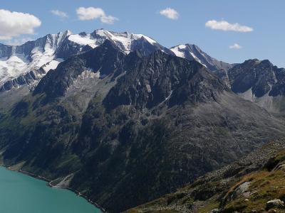 A high-angle landscape view of a turquoise alpine reservoir (Schlegeisspeicher) at the base of rugged, snow-capped mountain peaks in the Zillertal Alps.
