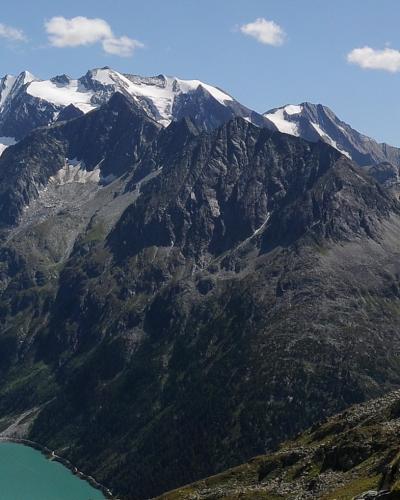 A high-angle landscape view of a turquoise alpine reservoir (Schlegeisspeicher) at the base of rugged, snow-capped mountain peaks in the Zillertal Alps.