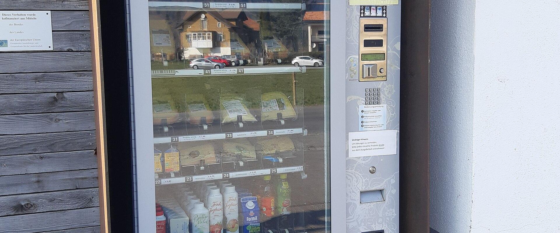 A white vending machine labeled "Produkt-Ausgabe" (Product Dispenser) containing dairy products like milk and cheese, housed in a wooden shelter with German signage including "Öffnungszeiten" (Opening Hours), set against a backdrop of alpine-style