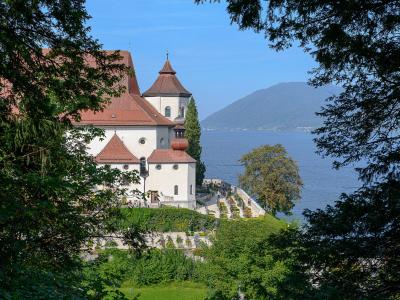 A white church with a red-tiled roof and a distinctive onion dome tower, situated on a grassy hill overlooking a large lake with mountains in the background, framed by trees in the foreground.
