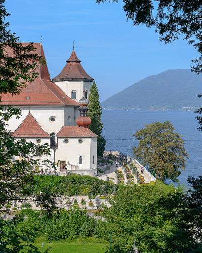 A white church with a red-tiled roof and a distinctive onion dome tower, situated on a grassy hill overlooking a large lake with mountains in the background, framed by trees in the foreground.