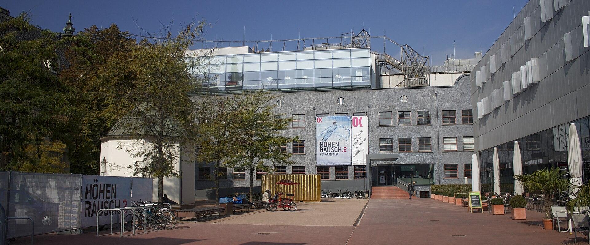 The image shows the exterior of the Offenes Kulturhaus Linz (OK), a contemporary art and culture center in Linz, Austria, with visible signage reading “HÖHEN RAUSCH 2” and the “OK” logo on banners and fencing.