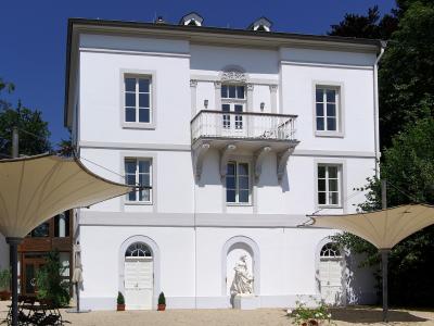 A white, three-story historic villa with a central balcony, arched ground-floor doorways, and a statue in a niche, set against a blue sky with trees and outdoor shade structures.