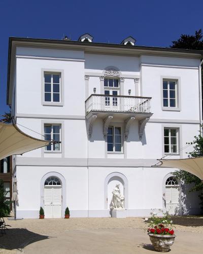 A white, three-story historic villa with a central balcony, arched ground-floor doorways, and a statue in a niche, set against a blue sky with trees and outdoor shade structures.