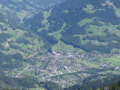 A high-angle panoramic view of a town nestled in a green alpine valley, showing dense housing, roads, and forested slopes.