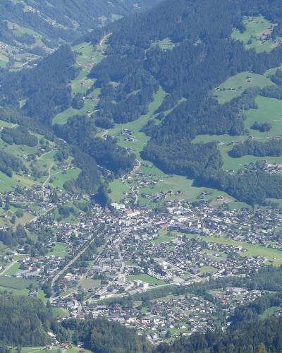 A high-angle panoramic view of a town nestled in a green alpine valley, showing dense housing, roads, and forested slopes.
