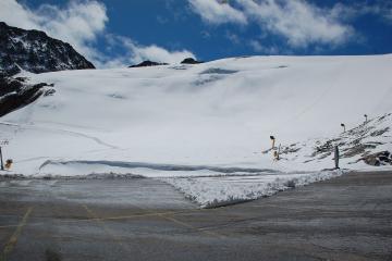 A high-altitude glacier slope with snowmaking equipment and a paved area in the foreground, under a blue sky with scattered clouds.