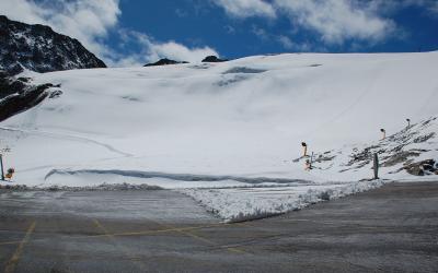 A high-altitude glacier slope with snowmaking equipment and a paved area in the foreground, under a blue sky with scattered clouds.