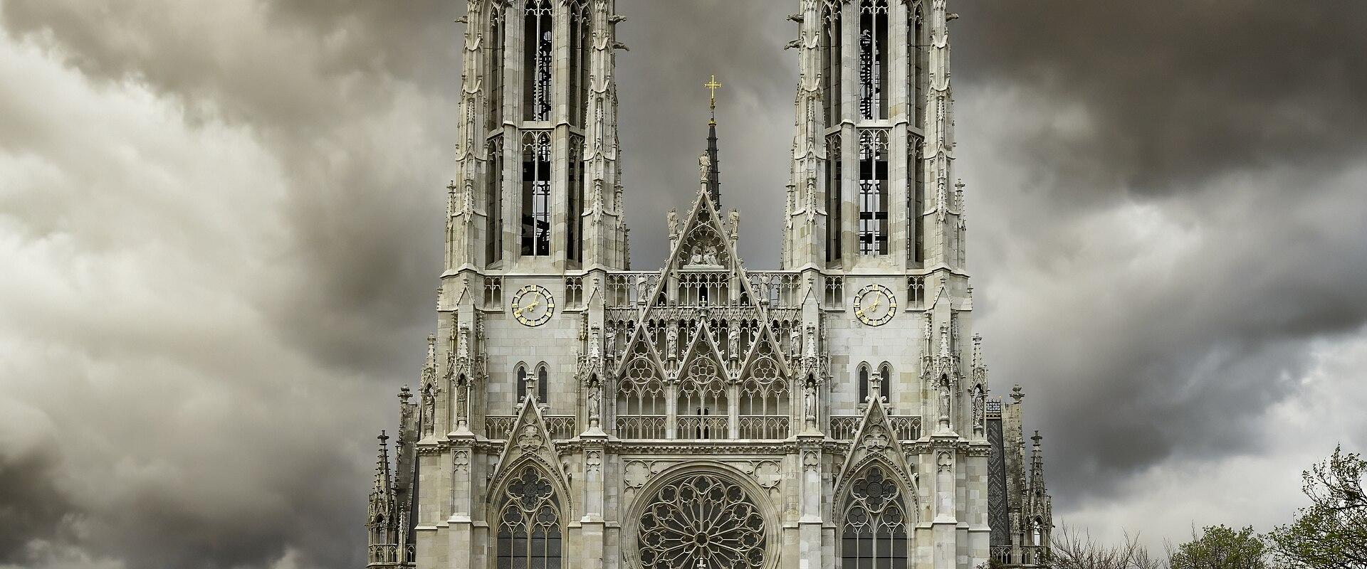 The image shows the Votivkirche (Votive Church), a 19th-century Neo-Gothic church in Vienna, Austria, featuring twin spires, a large rose window, and ornate stonework, set against a dramatic cloudy sky with green lawn and trees in the foreground.
