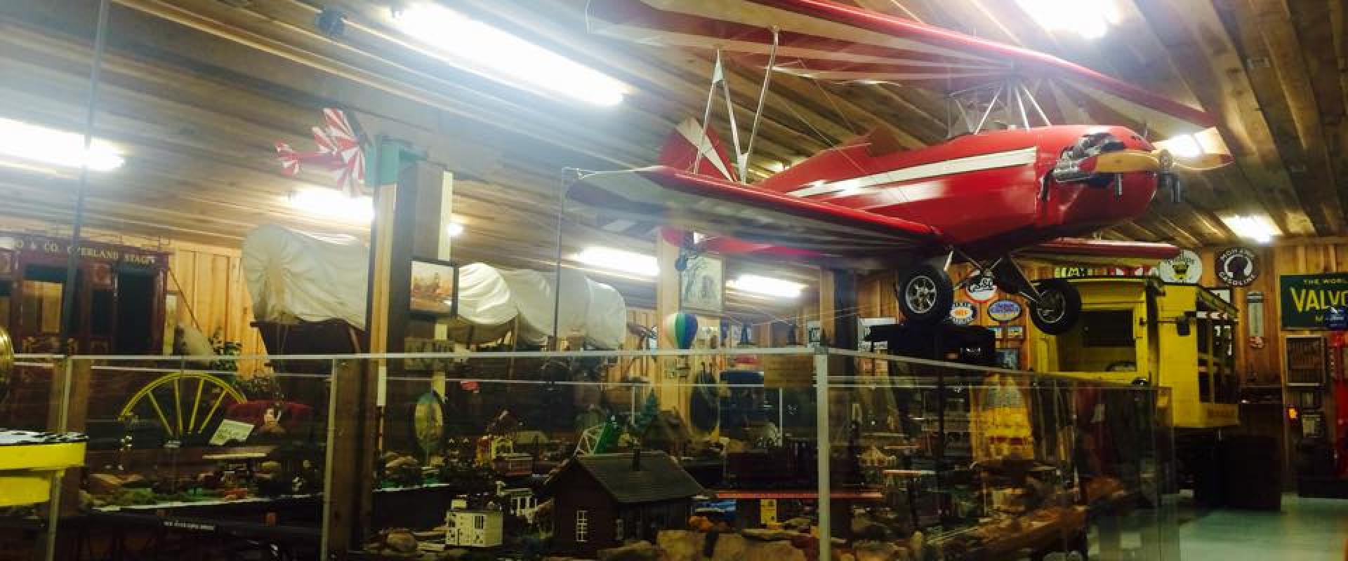 The image shows the interior of a museum with a red biplane suspended from a wooden ceiling, glass display cases containing model trains and other artifacts, and vintage signage including a "VALVO" sign and a covered wagon.