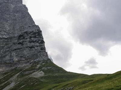 A steep, grey limestone mountain peak rises above a green alpine meadow under a cloudy sky, with a dirt path visible in the foreground.