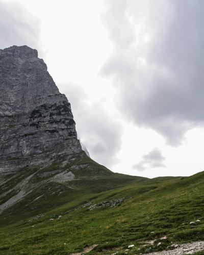 A steep, grey limestone mountain peak rises above a green alpine meadow under a cloudy sky, with a dirt path visible in the foreground.