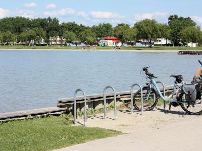 A view of a paved promenade along a lake shore with bicycles parked at metal racks, a wooden dock, and a grassy area with trees and buildings in the background.