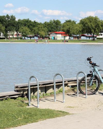 A view of a paved promenade along a lake shore with bicycles parked at metal racks, a wooden dock, and a grassy area with trees and buildings in the background.