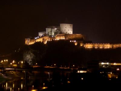 A night photograph of a large, illuminated fortress complex situated on a rocky hill overlooking a river and a town with streetlights.