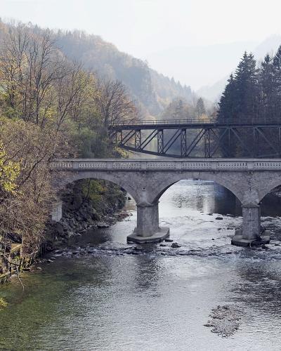 A stone arch bridge carrying a road crosses a river, with a metal truss railway bridge situated directly above it in a forested valley.