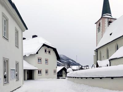 A view of a white-walled church building with a slate roof, a cemetery wall in the foreground, a wooden crucifix, and a distinctive jagged mountain peak in the background.