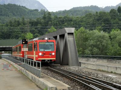 A red and cream narrow-gauge train with "ZILLERTALBAHN" written on the front and "Mayrhofen" on its destination display, traveling on tracks through a green, mountainous landscape near a modern concrete structure.
