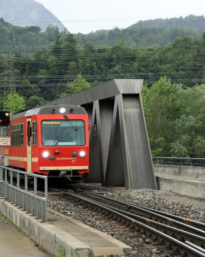 A red and cream narrow-gauge train with "ZILLERTALBAHN" written on the front and "Mayrhofen" on its destination display, traveling on tracks through a green, mountainous landscape near a modern concrete structure.