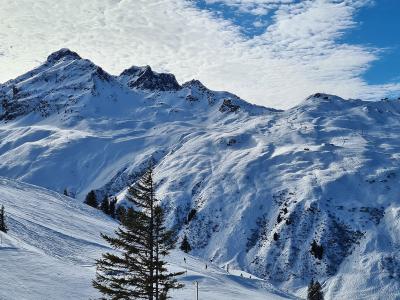 A panoramic view of a snow-covered mountain range with ski slopes, scattered pine trees, and visible ski lift infrastructure under a blue sky with clouds.
