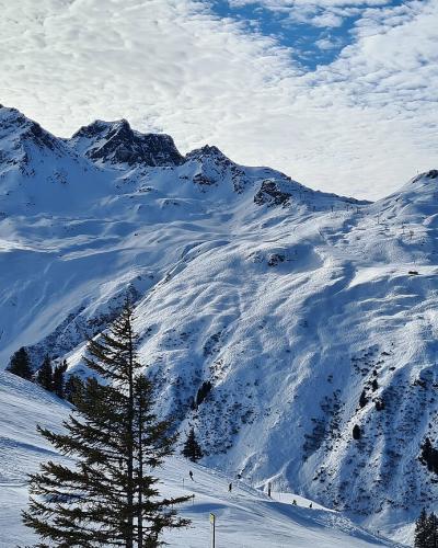 A panoramic view of a snow-covered mountain range with ski slopes, scattered pine trees, and visible ski lift infrastructure under a blue sky with clouds.