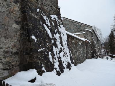 A low-angle view of a tall, dark stone fortification wall covered in patches of snow, with a snow-covered path running alongside it and a building with a sloping roof visible in the background.