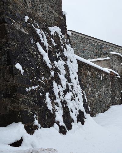 A low-angle view of a tall, dark stone fortification wall covered in patches of snow, with a snow-covered path running alongside it and a building with a sloping roof visible in the background.