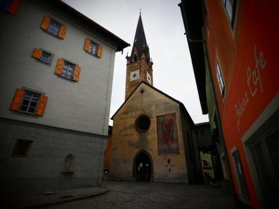 A low-angle view of a narrow cobblestone street in a European old town, featuring a church with a tall spire and a large mural on its facade, flanked by buildings with orange shutters and a sign for "Beluna Cafe".