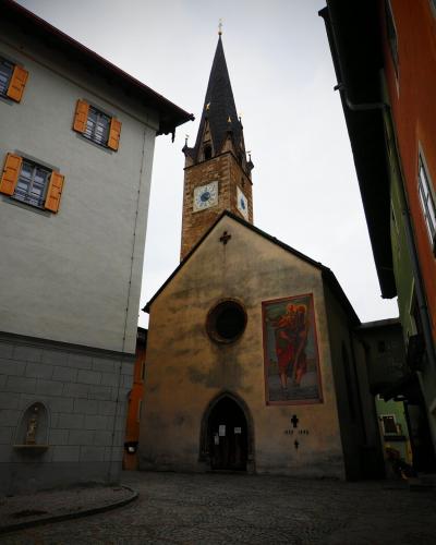 A low-angle view of a narrow cobblestone street in a European old town, featuring a church with a tall spire and a large mural on its facade, flanked by buildings with orange shutters and a sign for "Beluna Cafe".