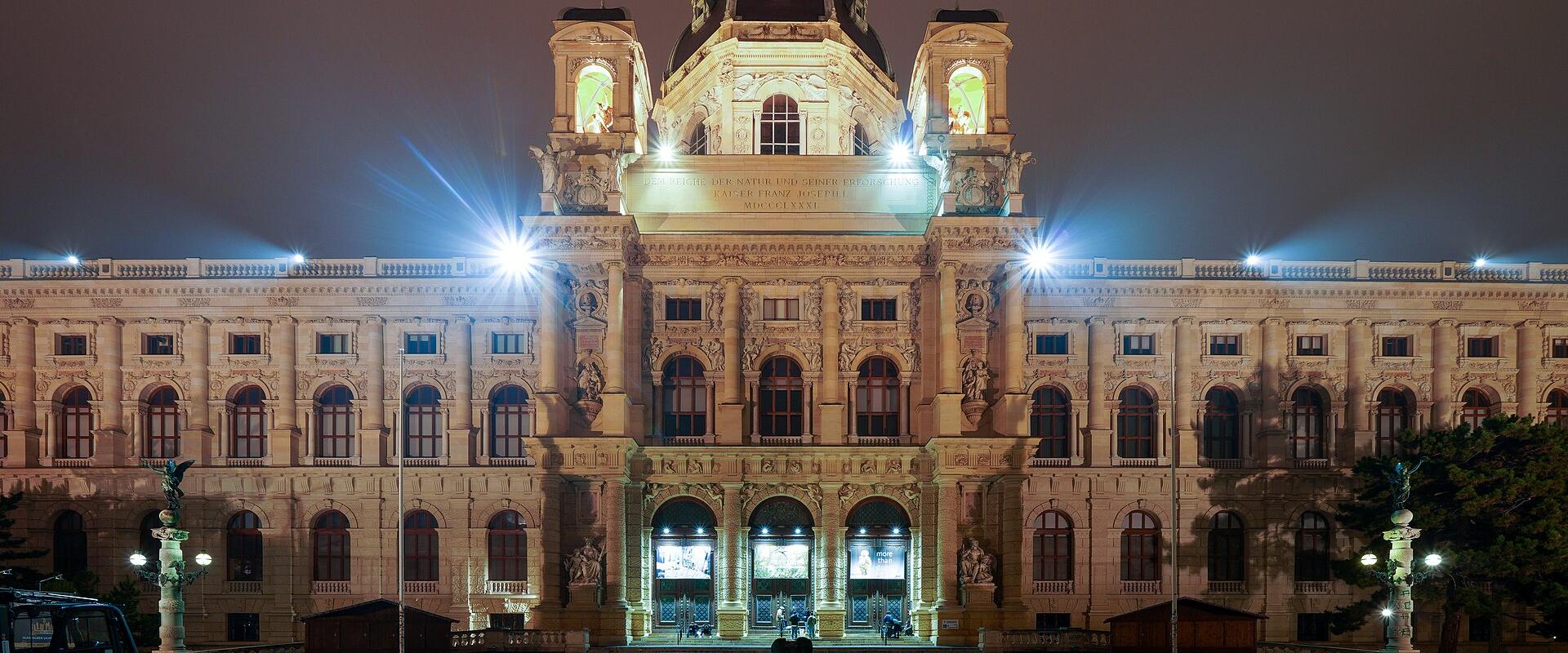 A grand, ornate neoclassical museum building illuminated at night, featuring a central dome with a statue on top, flanked by towers, and an inscription above the entrance reading “KUNSTHISTORISCHES MUSEUM WIEN” — clearly identifying it as the Kuns