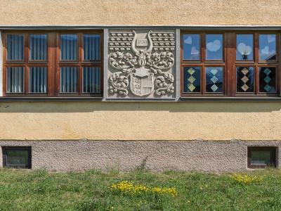 A close-up of a building facade featuring a large stone relief carving of a coat of arms with the date 1958, flanked by wooden-framed windows with decorative glass, set above a grassy slope.