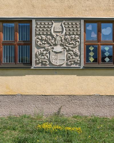 A close-up of a building facade featuring a large stone relief carving of a coat of arms with the date 1958, flanked by wooden-framed windows with decorative glass, set above a grassy slope.