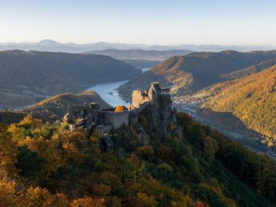A high-angle landscape photograph showing the stone ruins of a castle perched on a rocky cliff overlooking a wide river valley with autumn foliage and distant mountains.