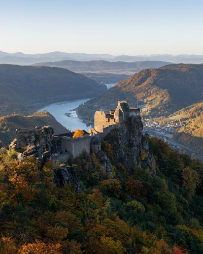 A high-angle landscape photograph showing the stone ruins of a castle perched on a rocky cliff overlooking a wide river valley with autumn foliage and distant mountains.