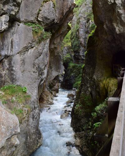 A view of a narrow, rocky gorge with a rushing stream, featuring a wooden walkway and tunnel entrance on the right side.